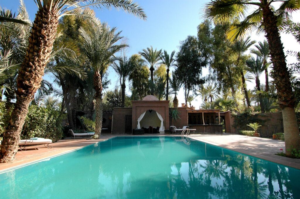 Opulent-Mediterra-Morocco-Luxury-Pool-Riad A luxury Moroccan riad pool surrounded by palm trees and traditional architecture, offering a serene atmosphere for curated cultural tours.
