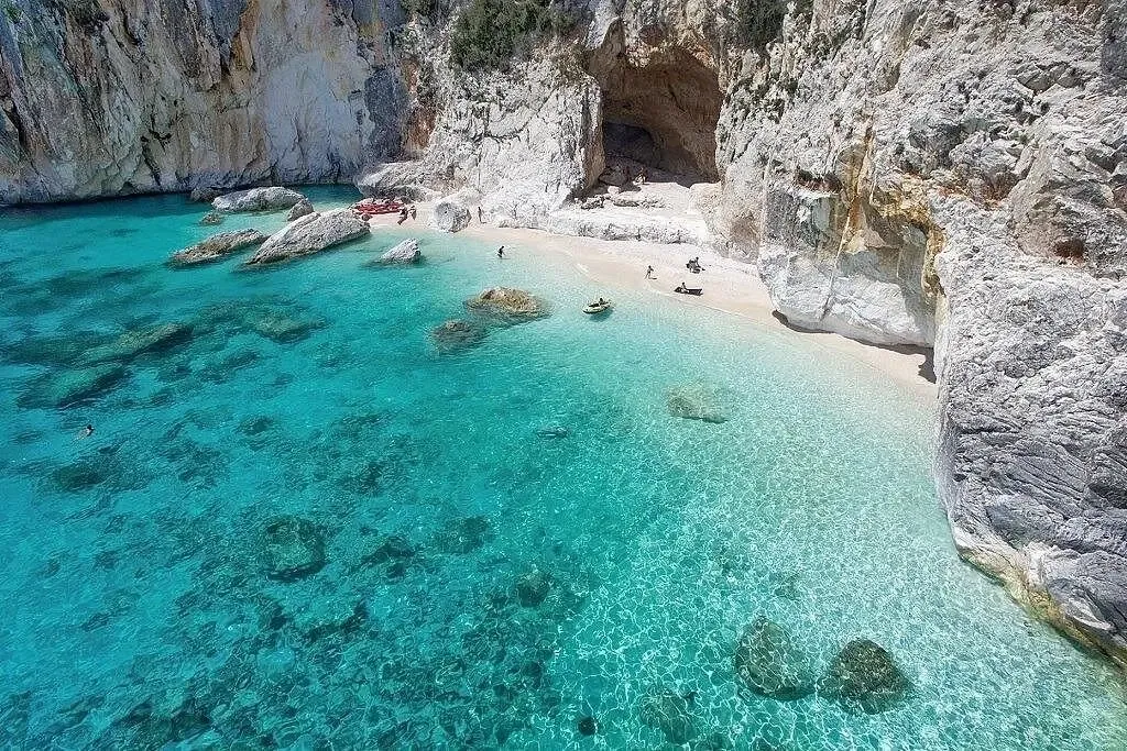 High-angle view of a stunning secluded cove beach in Sardinia with remarkably clear turquoise water and massive limestone cliffs. A natural sea cave frames the pebble shore, where a few kayaks and people are resting.