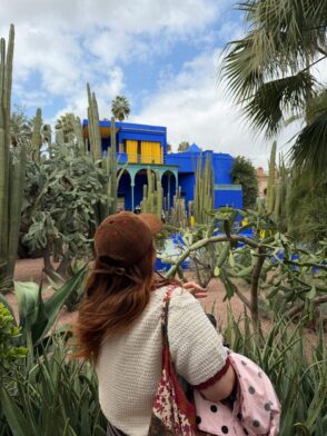 Cobalt & Cactus: A Quiet Moment in Jardin Majorelle A traveller with auburn hair and a brown hat looks at the iconic cobalt-blue architecture of the Jardin Majorelle in Marrakech, surrounded by cactuses and palm trees.