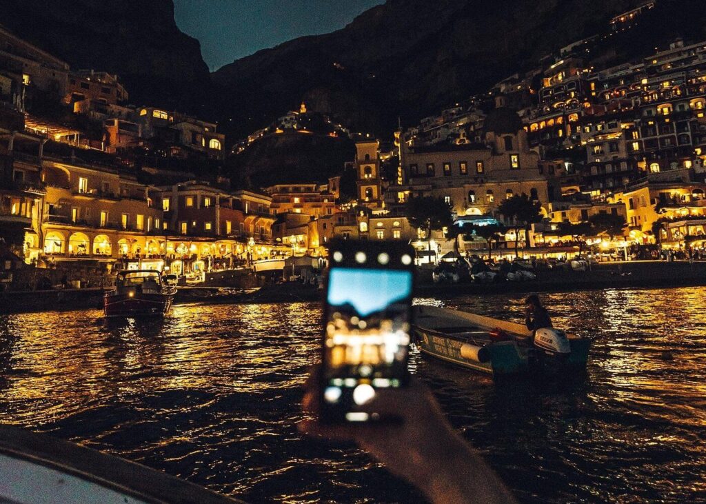 Midnight Gold: Capturing Positano’s Enchanting Nightscape A person holds up a smartphone to photograph the glittering cliffside village of Positano at night from a boat on the shimmering dark water.