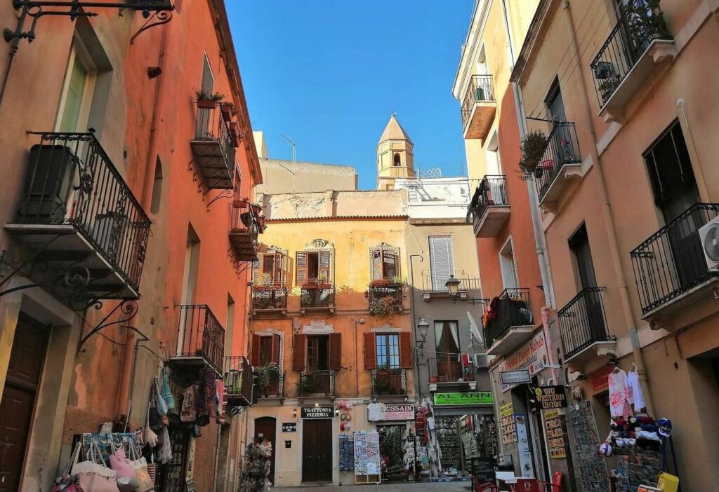 Cobblestones & Cannonau: Wandering Cagliari’s Historic Heart A vibrant, narrow street in Cagliari's historic old quarter lined with warm terracotta and pastel buildings, wrought-iron balconies, small local shops, and a historic church bell tower peeking into a clear blue sky.
