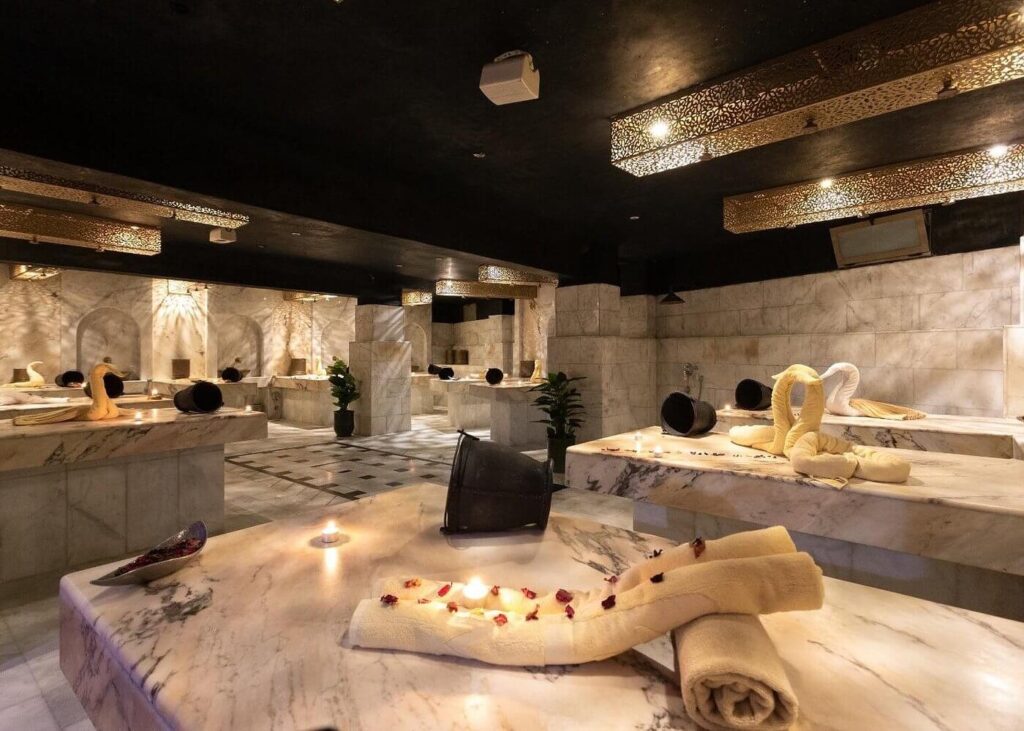 Sanctuary of Silk & Steam: The Moroccan Hammam Ritual A luxurious Moroccan hammam spa room featuring polished white marble treatment tables decorated with glowing candles, rose petals, and folded towels beneath ornate brass light fixtures.