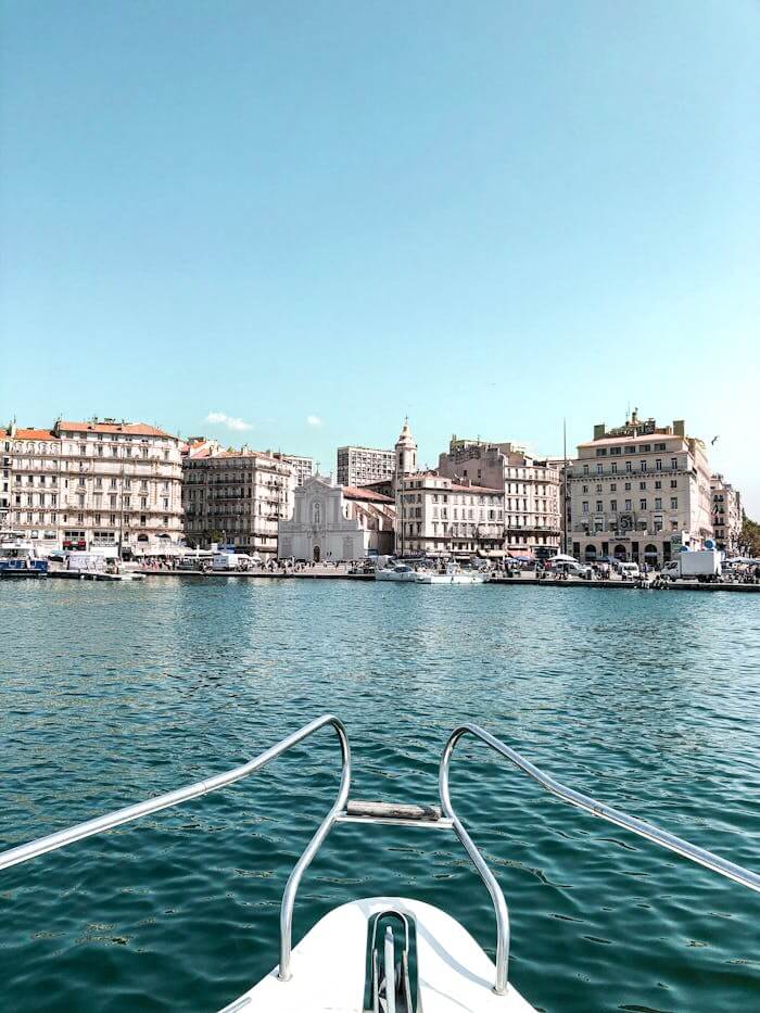 Opulent Mediterranean Harbor Experience | Luxury Coastal Boat View Luxury boat view of an opulent Mediterranean harbor, showing a white bow with metal handrails framing turquoise water and a historic waterfront with classical stone buildings and a domed church tower under a clear sunny sky.